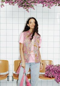 Un gilet floral rose sur un t-shirt rose clair, associé à un jean bleu clair. Tenant un sac rose, le tout devant un fond carrelé orné de fleurs.