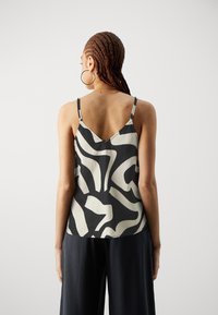 Camisole noir et crème à motifs avec des fines bretelles, présentant un dos en V et une texture lisse, porté avec un pantalon large sombre.