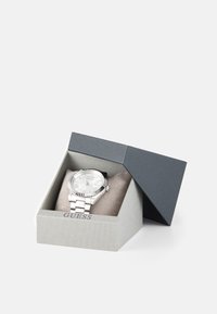 Sudraba pulkstenis ar nerūsējošā tērauda siksniņu, izvietots pelēkā kastē ar strukturētu apdari un mīkstu, bēša krāsas spilventiņu iekšpusē.