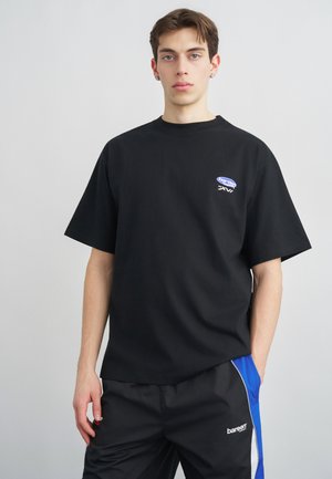 Jeune homme portant un t-shirt noir avec un petit logo et un pantalon noir à rayures bleues et blanches, debout devant un fond uni clair.