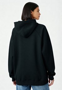 Sweat à capuche noir surdimensionné avec capuche à cordon, poignets et ourlet côtelés, et une texture lisse, associé à un jean en denim bleu clair.