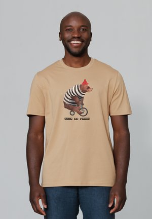 T-shirt beige avec un graphique d'un ours portant une chemise rayée et un bonnet rouge, faisant du vélo, avec le texte "EDGAR DE FRANZ" en dessous.