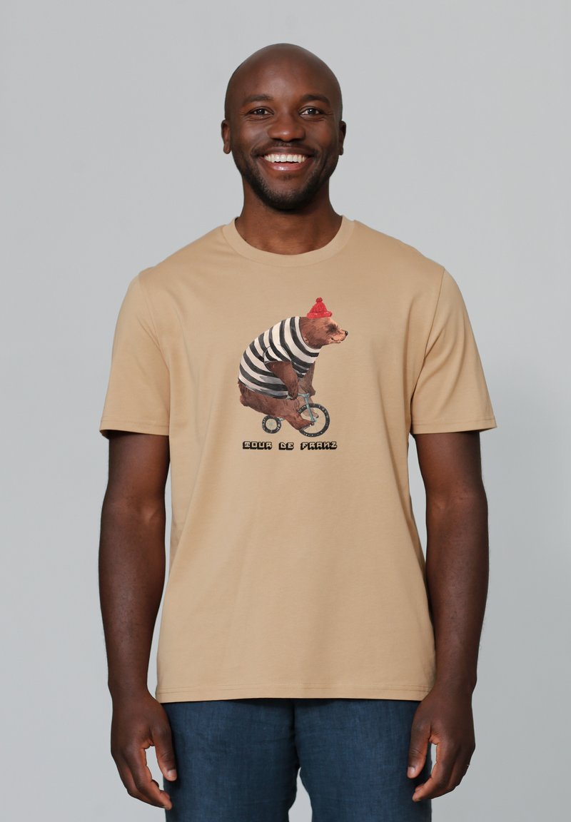 Tan t-shirt met een grafiek van een beer in een gestreepte shirt en een rode beanie, die op een fiets rijdt, met de tekst "EDGAR DE FRANZ" eronder.