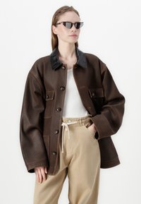 Brun skinnjakke med svart krage, med store knapper og frontlommer. Kombinert med beige bukser med snøring og en hvit topp.