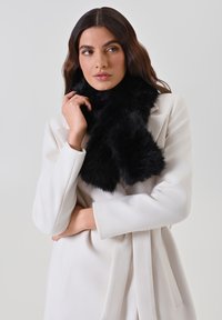 Écharpe en fausse fourrure noire avec une texture douce et un design volumineux, drapée de manière élégante autour du cou, associée à un manteau blanc.