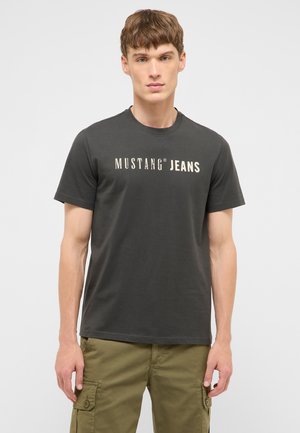 Junger Mann trägt ein dunkelgraues "Mustang Jeans" T-Shirt und olivgrüne Cargohosen, steht vor einem schlichten hellen Hintergrund.