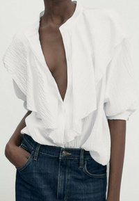 Blouse blanche texturée avec un décolleté en V profond et des détails volantés évasés, associée à un jean en denim bleu foncé. Manches bouffantes et coupe décontractée.