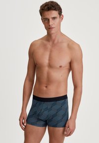 Blauwe boxer shorts met een patroon en een zwarte elastische tailleband. Het ontwerp toont geometrische vormen in een lichter blauw tegen een donkere achtergrond.