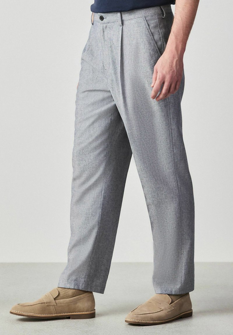 Homme portant un pantalon gris clair sur mesure et des mocassins en daim beige, debout sur un sol uni devant un fond clair.