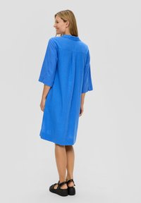 Robe en lin bleu à manches courtes avec une coupe décontractée, présentant un empiècement au dos et une longueur genou. Portée avec des sandales à plateforme noires.