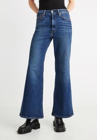 Τζιν παντελόνι καμπάνα σε σκούρο μπλε, με ψηλή μέση, φαρδύ ποδόγυρο και διακριτικές ραφές. Φορεμένο με μαύρα δερμάτινα παπούτσια πλατφόρμα.