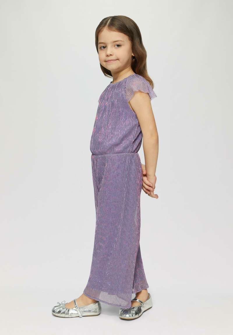 Fioletowy kombinezon z fakturowanej tkaniny, krótki, falbaniasty rękaw i elastyczny pas. Połączony z srebrnymi balerinami i jasnym tłem.