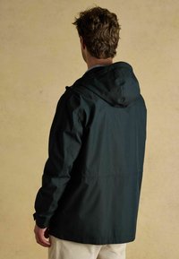Jachetă impermeabilă de culoare verde închis, cu kapuc, fermoar frontal și buzunare laterale, având o textură netedă și o croială lejeră. Vedere din spate.