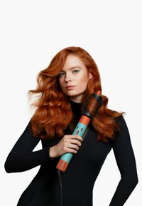 Rund hårborste med ett turkosa handtag och orange detaljer, med ett svart ventilerat fat. Modellen håller borsten med vågigt röd hår.