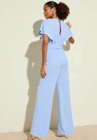 Jumpsuit bleu clair avec des manches à volants et un détail de trou clé au dos. Présente des jambes larges et une taille ceinturée pour une structure. Texture de tissu lisse.