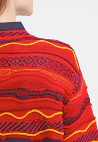 Červený pletený kardigan s modrými a žlutými akcenty, s vlnitými a geometrickými texturami. Detailní prošití po okrajích.