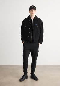 Schwarze Cordjacke mit Druckknöpfen, schwarzer Zip-Up-Pullover darunter, schwarze Cargo-Hosen und schwarze Turnschuhe. Schwarze Mütze mit "BOSS"-Logo.