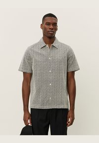 Chemise à manches courtes en tissu gris clair avec un motif paisley sombre, dotée d'une fermeture à boutons et d'un col évasé, portée avec un pantalon noir.