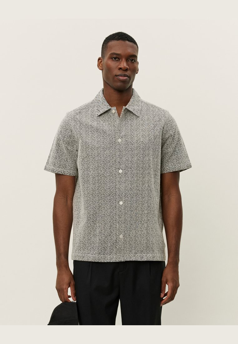 Chemise à manches courtes en tissu gris clair avec un motif paisley sombre, dotée d'une fermeture à boutons et d'un col évasé, portée avec un pantalon noir.