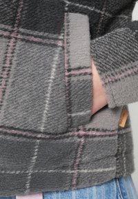 Pulover gri din fleece cu un model în carouri de nuanțe deschise și închise de gri, accentuat cu linii roz. Are un buzunar lateral și o etichetă cu brand.