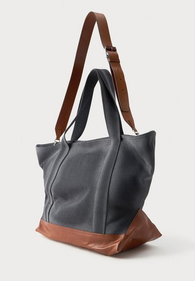 Tamsiai pilkas drobės krepšys su rudu odiniu dugnu, dviem rankenomis ir reguliuojamu diržu. Yra struktūrizuotas dizainas ir minkšta tekstūra.