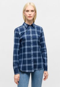 Woman wearing navy blue plaid long-sleeve button-up shirt and light blue jeans, standing against a plain light background.