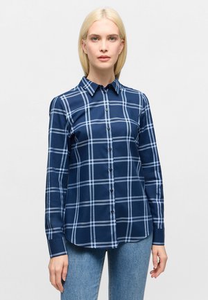 Woman wearing navy blue plaid long-sleeve button-up shirt and light blue jeans, standing against a plain light background.