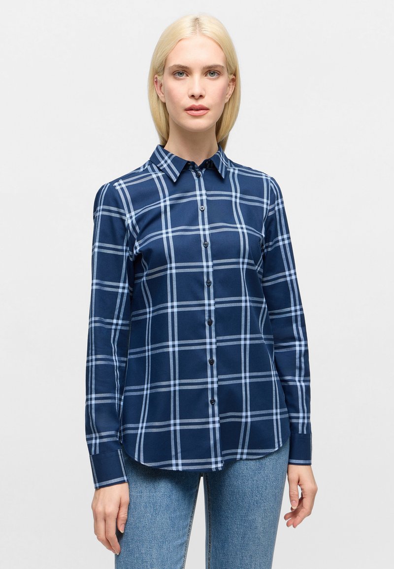 Woman wearing navy blue plaid long-sleeve button-up shirt and light blue jeans, standing against a plain light background.