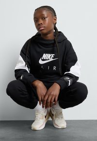 Zwarte Nike Air hoodie met witte accenten en logo, gecombineerd met zwarte sweatpants en crèmekleurige Nike sneakers met kastanjebruine details.