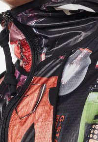 Veste à capuche colorée avec des imprimés graphiques. Elle est équipée d'une fermeture éclair noire, d'un cordon de serrage et d'un tissu texturé. Les détails de design remarquables incluent des couleurs vives et des motifs audacieux.