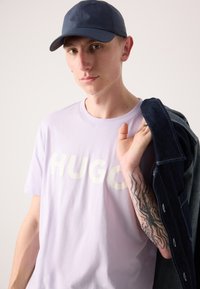 Light purple T-shirt with "HUGO" in cream lettering. A dark blue cap and navy denim jacket are draped over one shoulder. Features a tattooed arm.