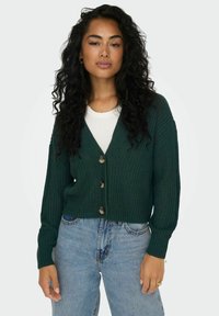 Cardigan lavorato a maglia verde scuro con scollo a V, cinque bottoni e polsini a coste, indossato sopra una maglietta bianca e jeans blu.