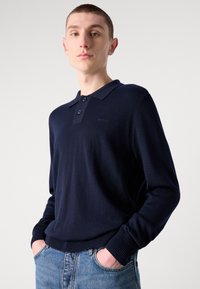 Sötétkék, hosszú ujjú póló kötött pulóver három gombbal és bordázott mandzsettával, a mellkason diszkrét logóhímzéssel.