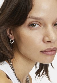 Zilveren hoepel oorbellen met een gedraaid ontwerp, voorzien van een gladde metallic afwerking. Draagt een model met donker haar en een zilveren ketting.