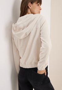 Lichtbeige corduroy hoodie met een ontspannen pasvorm, ribbeltextuur, trekkoordkap en elastische onderrand. Gecombineerd met zwarte broek.