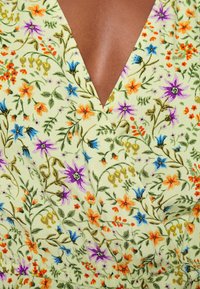 Robe florale jaune à encolure en V, présentant des fleurs orange, violettes et bleues avec des feuilles vertes sur un tissu jaune clair.