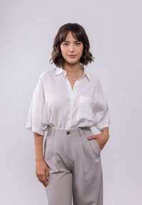 Weißes Kurzarmblusenshirt mit einer vorderen Tasche, kombiniert mit hoch taillierten, hellgrauen Hosen mit einem plissee Design.