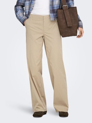 Persona, kas valkā bēšas krāsas platu bikšu modeli, baltu augšdaļu, zilu pleda raksta kreklu, tur brūnu zamšādas rokassomu un tumšas mokasīna stila kurpes.