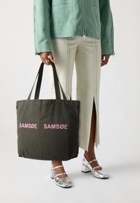 Mørkegrønn tote-veske med lyserødt "SAMSOE"-tekst, stoffmateriale, doble svarte stropper og en strukturert form. Modellen har på seg sølvfargede sko.