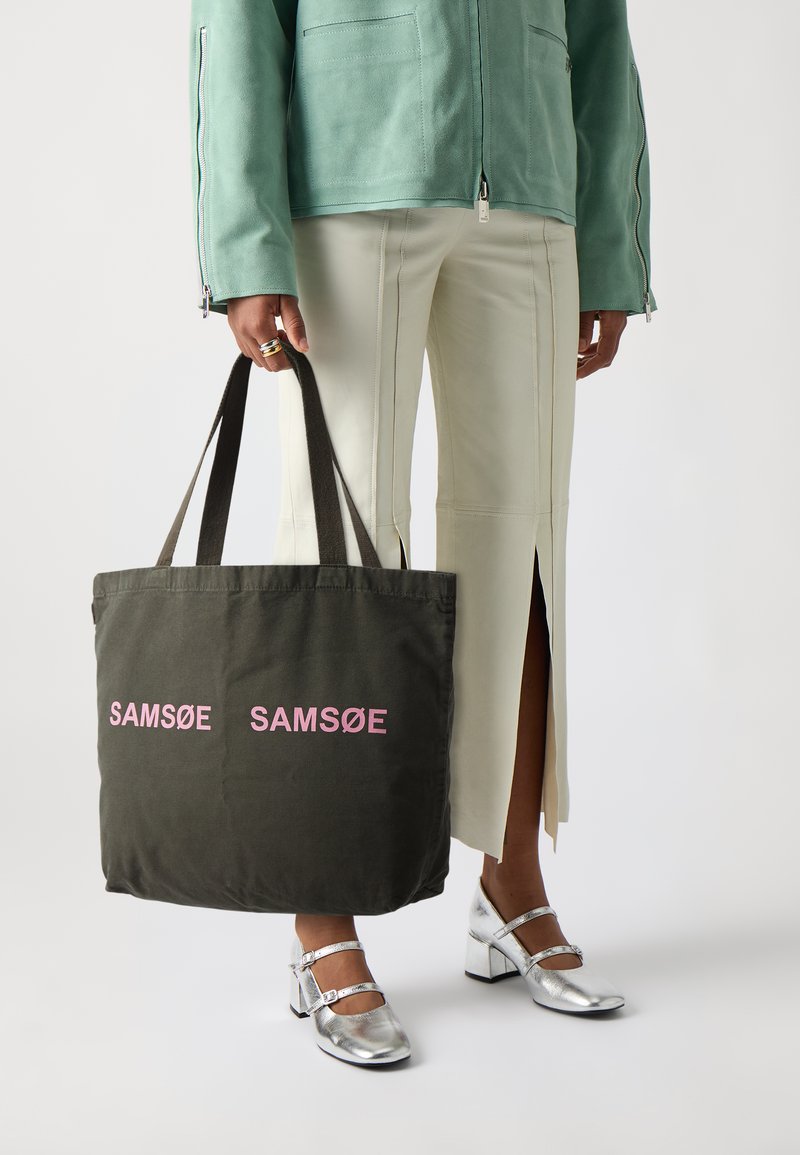 Mørkegrønn tote-veske med lyserødt "SAMSOE"-tekst, stoffmateriale, doble svarte stropper og en strukturert form. Modellen har på seg sølvfargede sko.