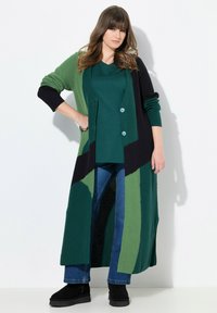 Langer gestrickter Cardigan in Grün und Schwarz, mit einem strukturierten Muster, zwei hellblauen Knöpfen, kombiniert mit einem grünen Oberteil und blauen Jeans.