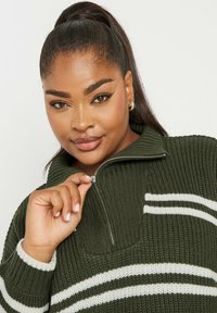 Stickad tröja i mörkgrön med vita ränder. Har en zippered krage, ribbad textur och en avslappnad passform.