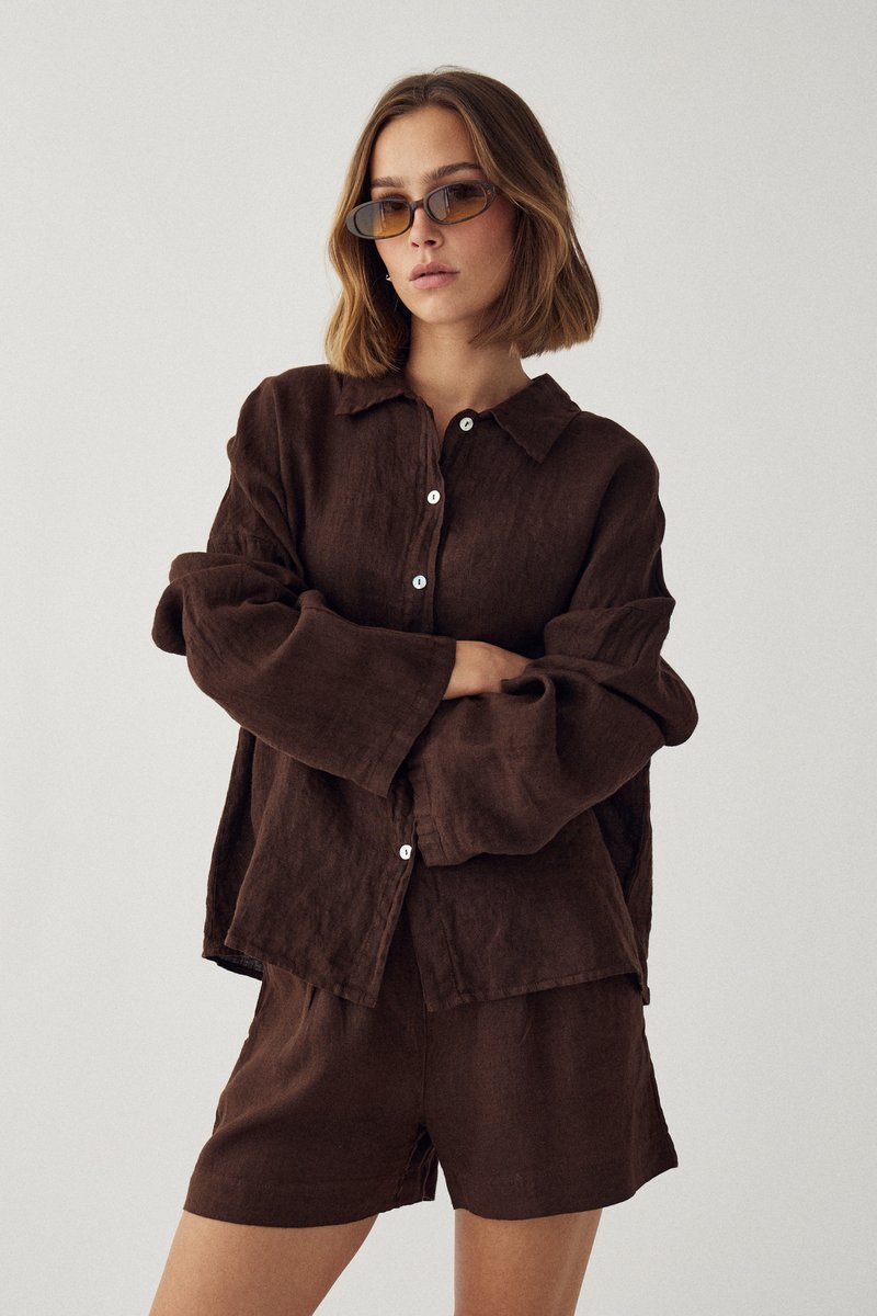 Femme portant une chemise marron à manches longues boutonnée et un short marron assorti, avec des cheveux courts et de petites lunettes de soleil teintées, les bras croisés.