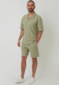 Hellgrünes gestricktes Kurzarmhemd mit Knopfverschluss und passenden Shorts. Das Model trägt weiße Sneaker. Strukturiertes Gewebe mit einem dezenten Muster.