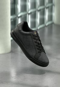 Schwarzer Ledersneaker mit Schnürsenkeln, der über einem polierten Betonboden in der Nähe einer Glasbausteinwand und einer weißen Säule schwebt.