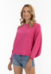 Vaaleanpunainen pulloveri, jossa on pitkät, löysät hihat ja neulospinta. Yhdistetty farkkushortsien kanssa, joissa on risaiset helmat.