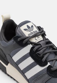 Grå och vit Adidas-sneaker med tre ränder, svarta mockadetaljer, vävd textur och knutna snören på en ljus bakgrund.