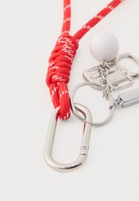 Cordon tressé rouge avec accents blancs, équipé d'un mousqueton en métal, d'un porte-clés et d'une petite sphère blanche. Comprend des pièces métalliques brillantes.