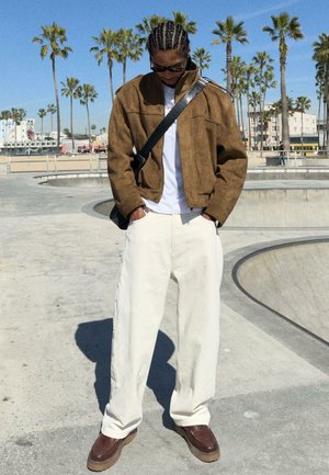 Mann mit geflochtenem Haar, der eine Sonnenbrille, eine braune Jacke, weiße Hose und braune Schuhe trägt und neben einem Skatepark steht, im Hintergrund Palmen.