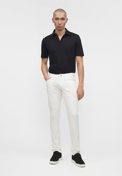 Άνδρας που στέκεται με το ένα χέρι στην τσέπη, φορά μαύρο κοντομάνικο πόλο, λευκό παντελόνι και μαύρα αθλητικά παπούτσια με απλό φόντο.
